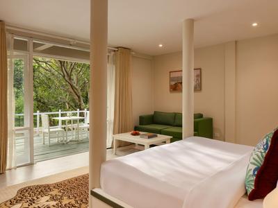 Interno di una camera d'hotel con letto, divano verde e balcone con vista sul verde.