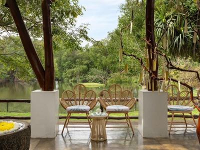 Zastřešená terasa se židlemi, stolem a výhledem na zelenou zahradu a rybník.