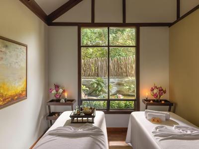 Světelná spa místnost se dvěma masážními lehátky, dekorativními květinami a velkým oknem s výhledem na zeleň.