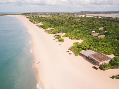 Rozsáhlá písečná pláž s hustou zelení a roztroušenými chatkami u moře pod zataženou oblohou.