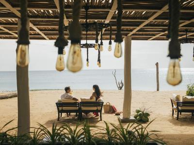 Dva lidé sedí u stolu pod zastřešenou terasou s výhledem na pláž a moře.