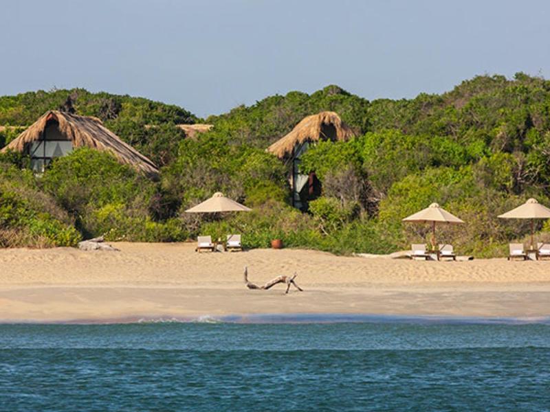 Spiaggia di sabbia con ombrelloni davanti a vegetazione verde e capanne sullo sfondo.