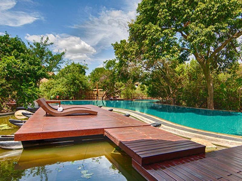 Piscina moderna con passerella in legno e lettini, circondata da alberi verdi e cielo blu.