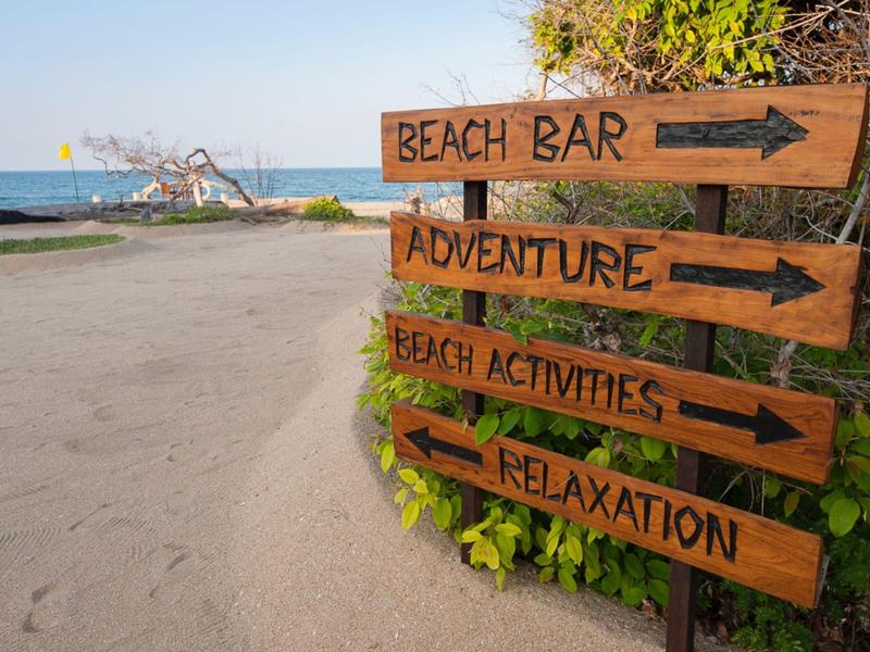 Cartello sulla spiaggia con indicazioni per beach bar, avventura, attività e relax.