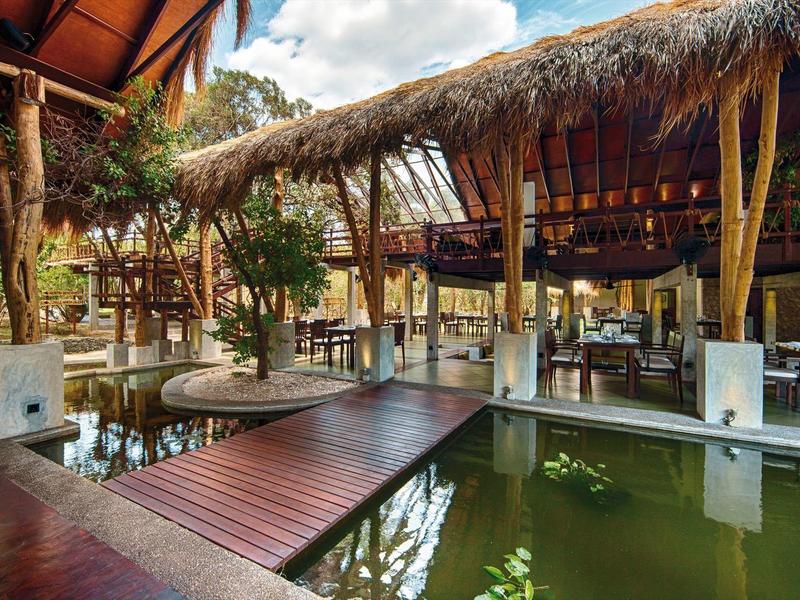 Ristorante all'aperto con passerella in legno sull'acqua e tetto di foglie di palma in ambiente naturale.