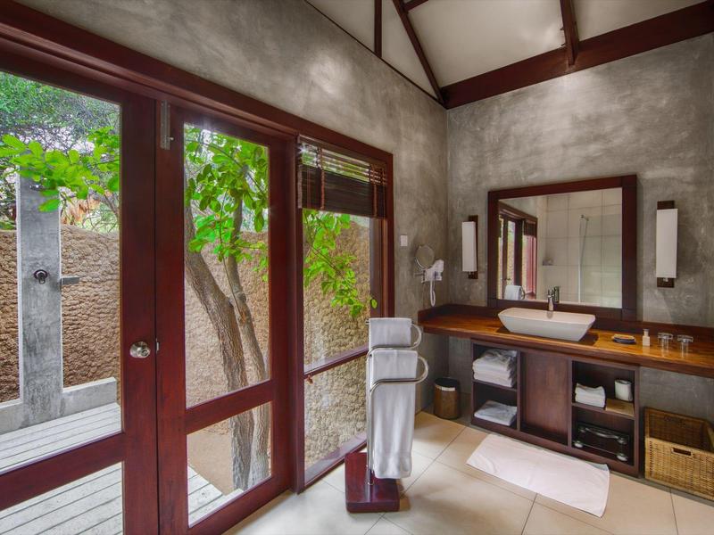 Bagno luminoso con grandi porte finestre, lavabo su tavolo di legno e vista su un'area esterna.