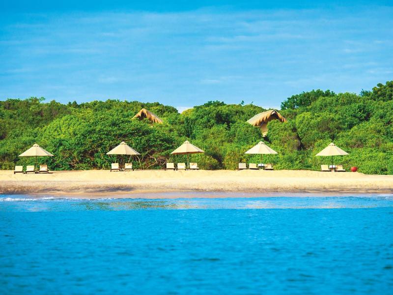Spiaggia con diversi padiglioni di paglia davanti a cespugli verdi e acqua blu limpida.