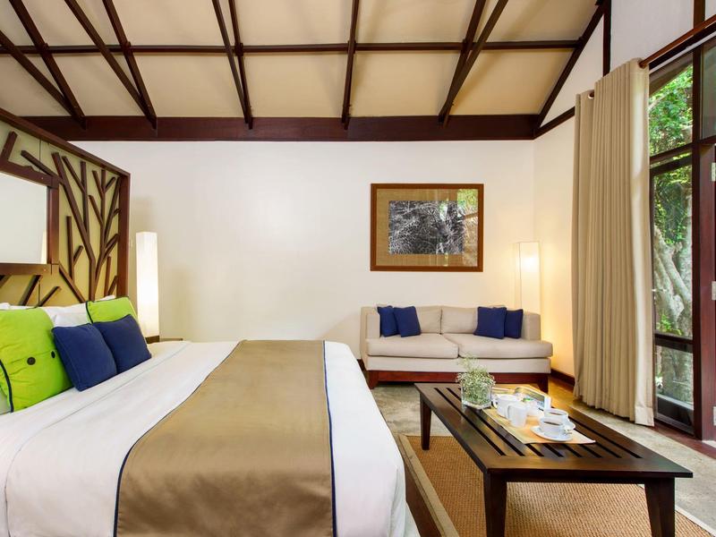 Camera d'hotel luminosa con letto grande, divano, soffitto in legno e vista sul giardino.