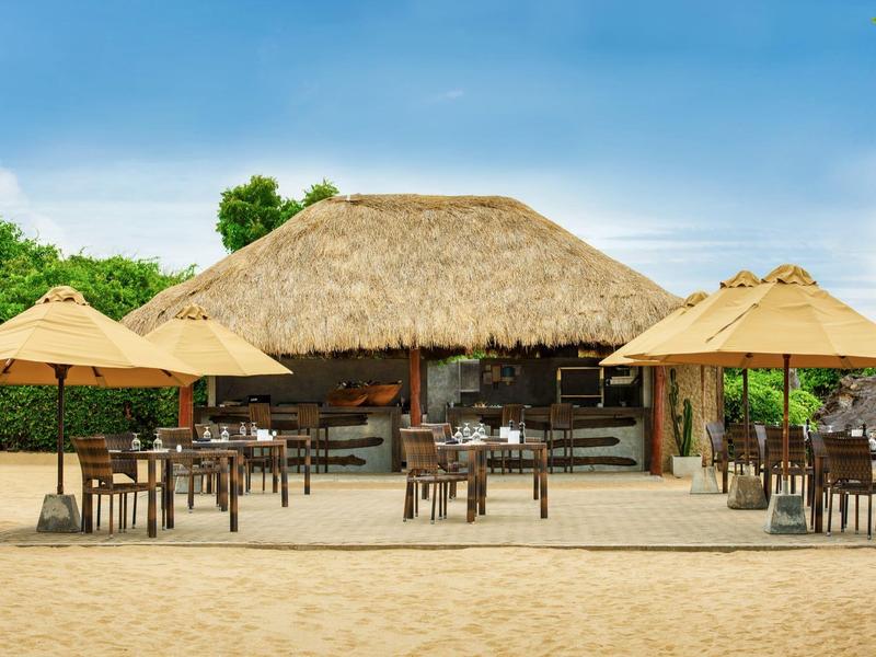 Bar sulla spiaggia con tetto in paglia, tavoli e ombrelloni sulla sabbia sotto un cielo blu.