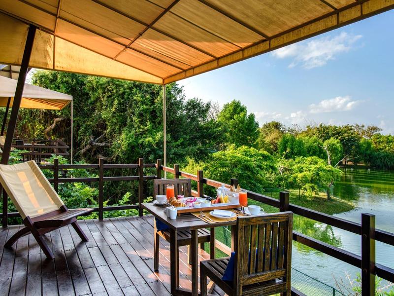 Balcone coperto con sedie di legno e tavolo con bevande accanto a un lago e natura verde.