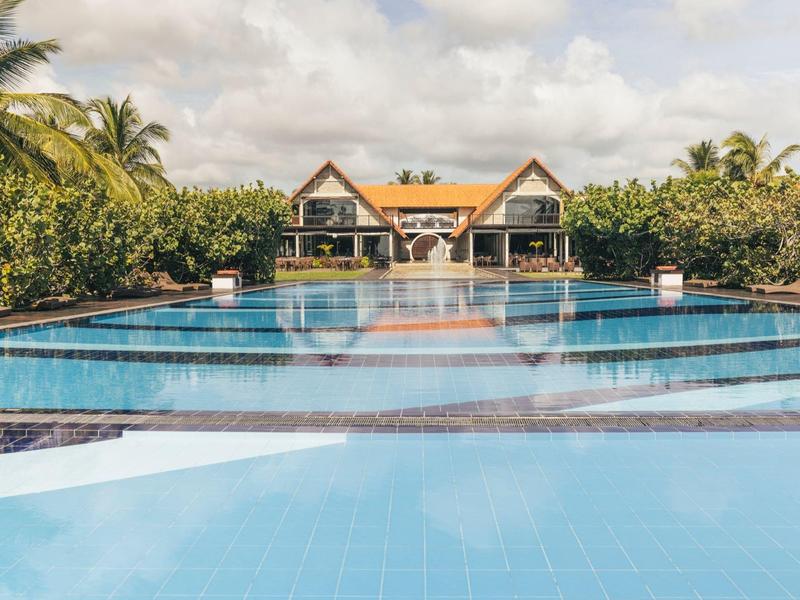 Grande piscine avec vue sur deux maisons et des palmiers sous un ciel nuageux.