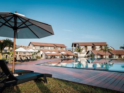 Großer Pool mit Holzterrasse, Liegestühlen und Sonnenschirmen vor gepflegten Gebäuden bei klarem Himmel.