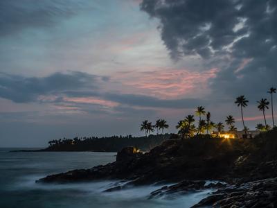 Dämmerung an einer Küste mit Palmen und Lichtern am Strand unter bewölktem Himmel.