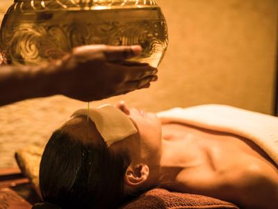 Person liegt entspannt, während warmes Öl im Wellnessbereich aufs Gesicht gegossen wird.