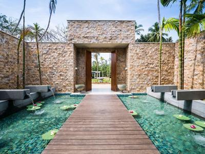 Holzsteg über einem Wasserbecken führt zu einem steinernen Tor in tropischer Umgebung.