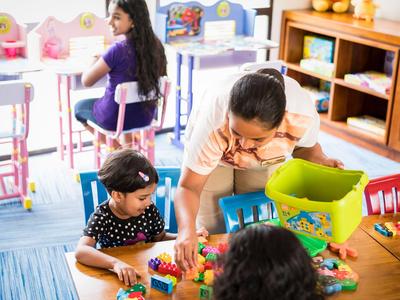 Kinder und Betreuerin bauen gemeinsam mit bunten Bauklötzen am Boden in einem Klassenraum.
