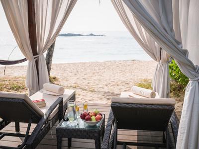 Zwei Liegestühle und ein Tisch mit Obst und Getränken unter einem weißen Vorhang am Strand.