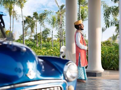 Ein Hotelträger in traditioneller Kleidung steht vor einer Säule neben einem blauen alten Auto.