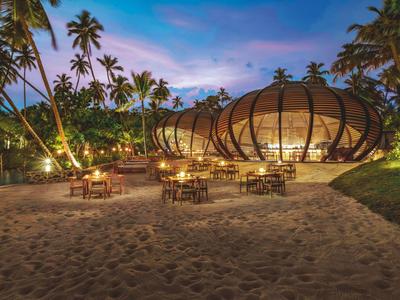 Abendlicher Strand mit beleuchteten, runden Strandhütten und Tischen unter Palmen.