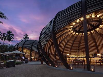 Abendaufnahme eines modernen, beleuchteten Strandrestaurants mit Palmen und lila Himmel.