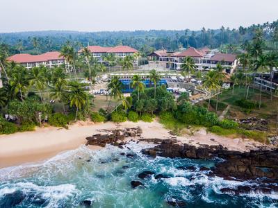 Hotelanlage mit Pool, umgeben von Palmen, direkt an einem felsigen und sandigen Strand.
