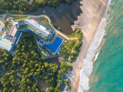 Vista aerea di un hotel con grandi piscine, circondato da vegetazione e spiaggia adiacente.