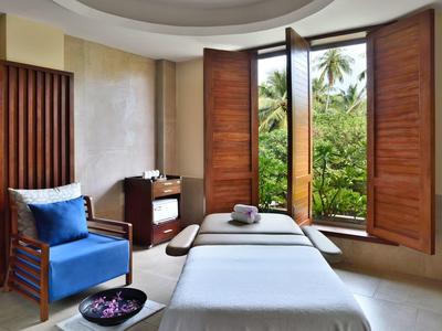 Helles Wellnesszimmer mit Massageliegen, blauem Sessel und offenem Fenster mit tropischem Ausblick.