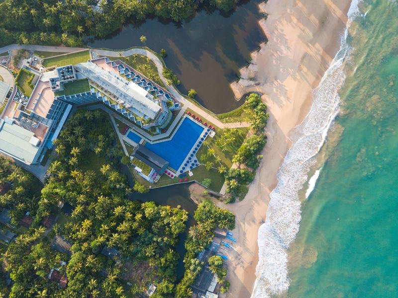 Vista aerea di un hotel con piscina, circondato da foresta e adiacente a una spiaggia sabbiosa e al mare.