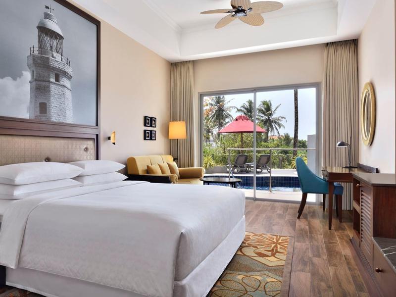 Camera d'hotel luminosa con letto grande, divano, scrivania e vista su un balcone con ombrellone rosso.