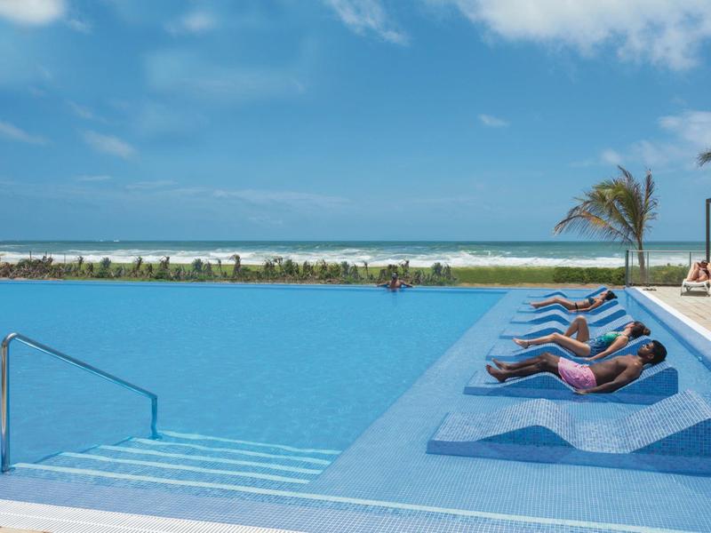 Piscina con lettini integrati di fronte al mare sotto un cielo azzurro.