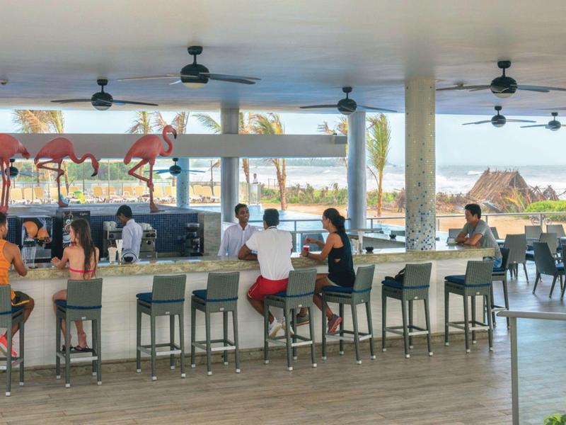 Bar all'aperto con sgabelli, ventilatori e vista sul mare con palme.