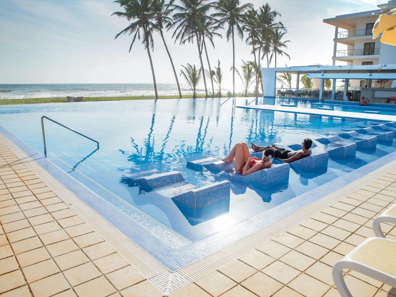 Piscina di un hotel con lettini integrati, palme e vista sull'oceano sullo sfondo.