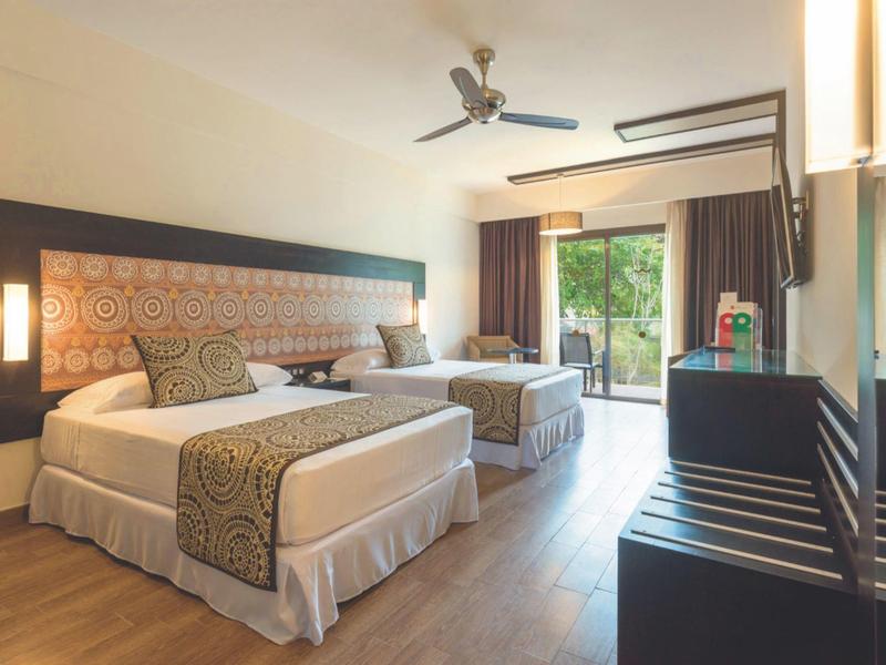 Camera d'hotel luminosa con due grandi letti, pavimento in legno e balcone verde.