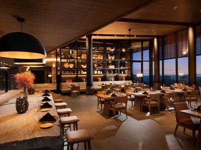 Moderní hotelová restaurace s dřevěným nábytkem, velkým oknem s večerním výhledem a elegantním osvětlením.