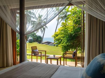 Blick aus einem Hotelzimmer auf eine Terrasse mit Stühlen und einem Tisch, umgeben von grünem Garten und Palmen.