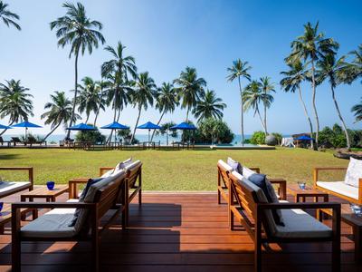 Outdoor-Lounge mit Holzboden und Sitzmöbeln vor einer Wiese mit Palmen und Sonnenschirmen am Meer.
