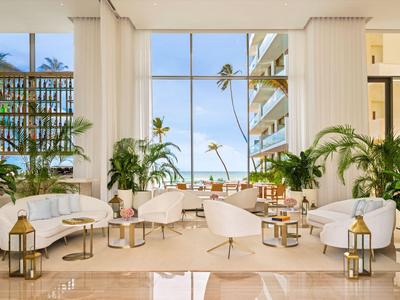 Lichte hotelkamer met wit meubilair en uitzicht op palmbomen en blauwe lucht.