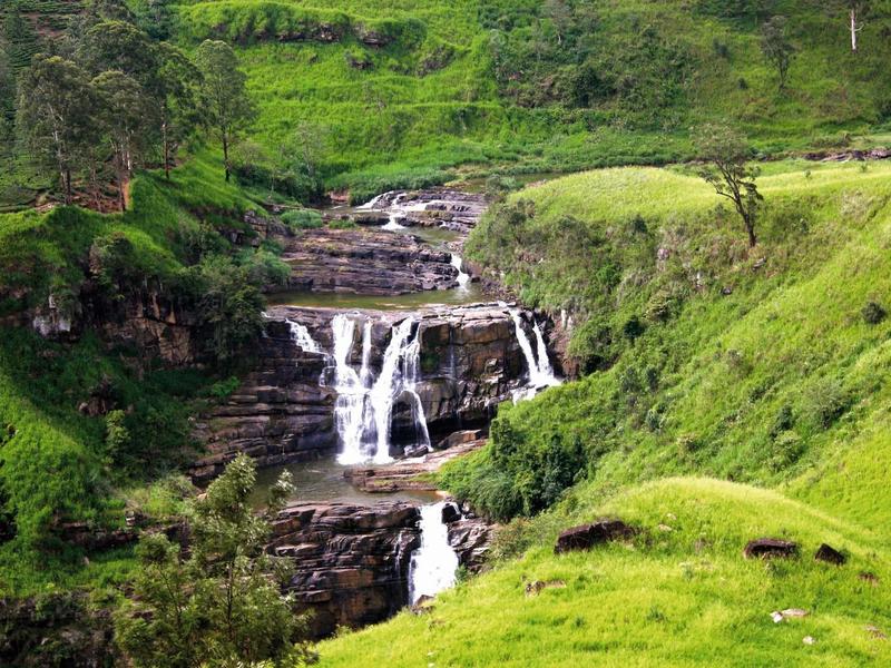 Grüner Hügel mit kleinem mehrstufigem Wasserfall und Bäumen im Hintergrund.