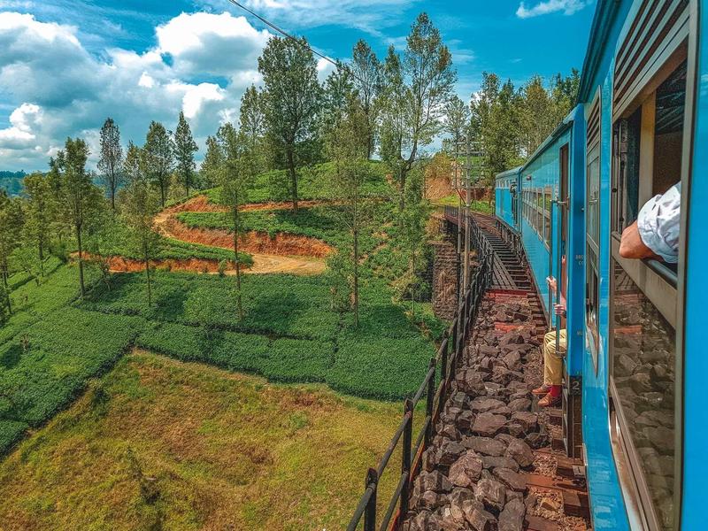 Blauer Zug fährt durch grüne Landschaft mit Bäumen und hügeligen Feldern unter blauem Himmel.