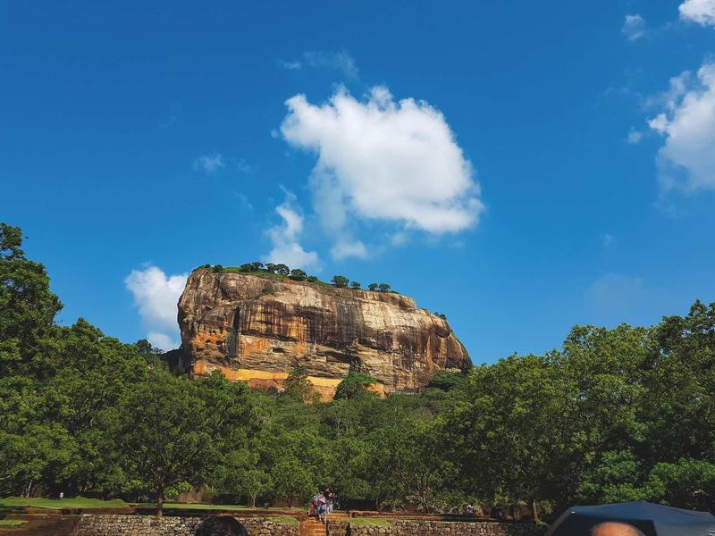 Großer Felsen mit glatter Oberfläche, umgeben von grünem Wald und blauem Himmel mit Wolken.