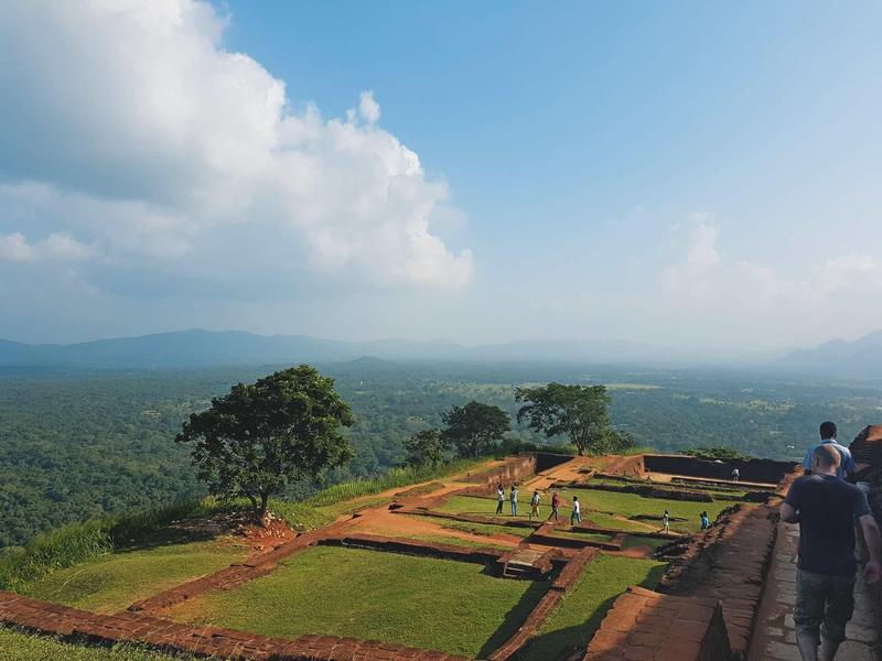 Berglandschaft mit grünem Wald, bewölktem Himmel und Menschen auf einem ausgedehnten Plateau.