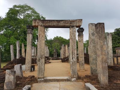 Alte steinerne Säulen und Torbogen in einer Ruine, im Hintergrund Bäume und grauer Himmel.