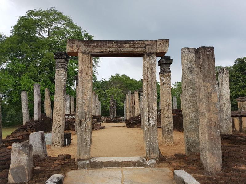 Starożytne ruiny z kamiennymi filarami i bramą, otoczone zielenią i pochmurnym niebem.