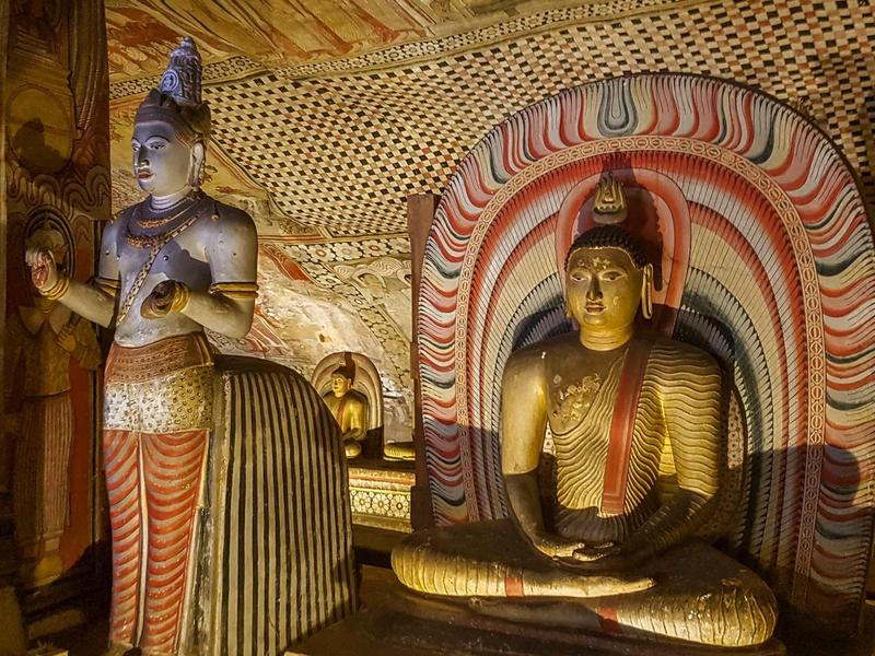 Innere eines Tempels mit sitzender Buddha-Statue und stehender Figur neben bemalten Wänden.