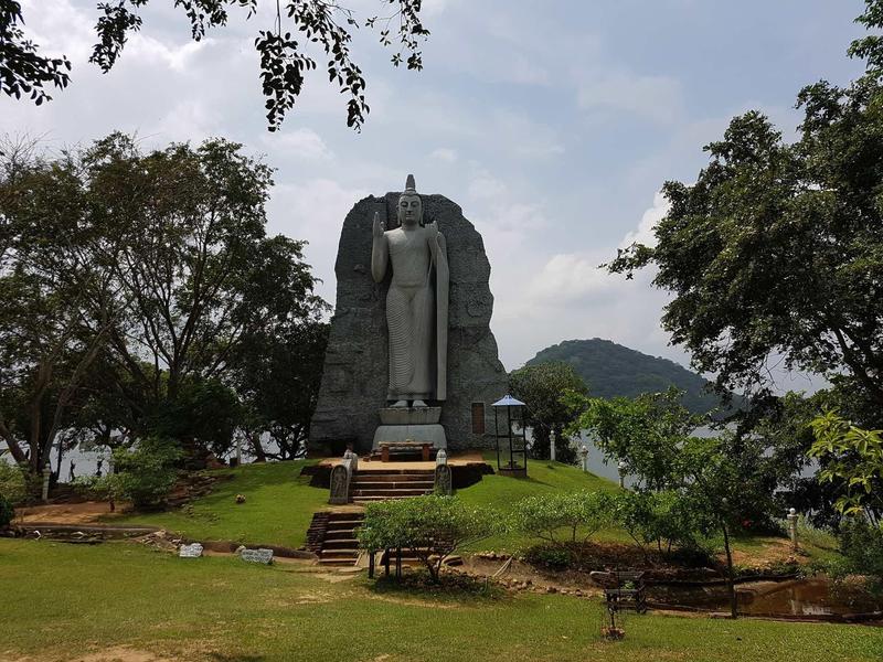 Große steinerne Buddha-Statue auf grünem Hügel, umgeben von Bäumen und Bergen im Hintergrund.