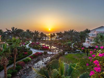 Sunset over a tropical garden with palm trees, flowers, small river, and resort buildings.