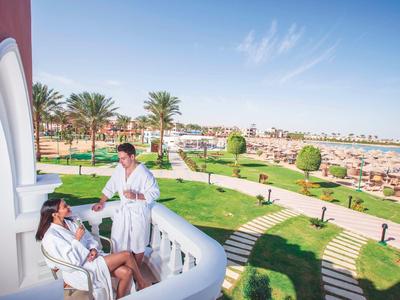Twee mensen in badjassen op een balkon met uitzicht op een resort met palmbomen en een strand.