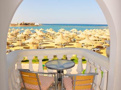 Balkonzicht door een boog met twee stoelen, tafel, parasols, zandstrand en blauwe zee.