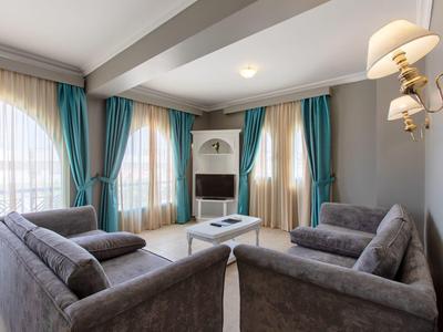 Gezellige hotelwoonkamer met grijze banken, turquoise gordijnen en een centrale witte salontafel.