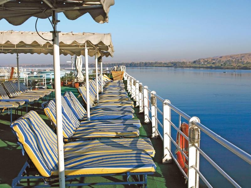 Lounge stoelen met kleurrijke kussens op een schip met uitzicht op een rustige rivier onder een blauwe lucht.